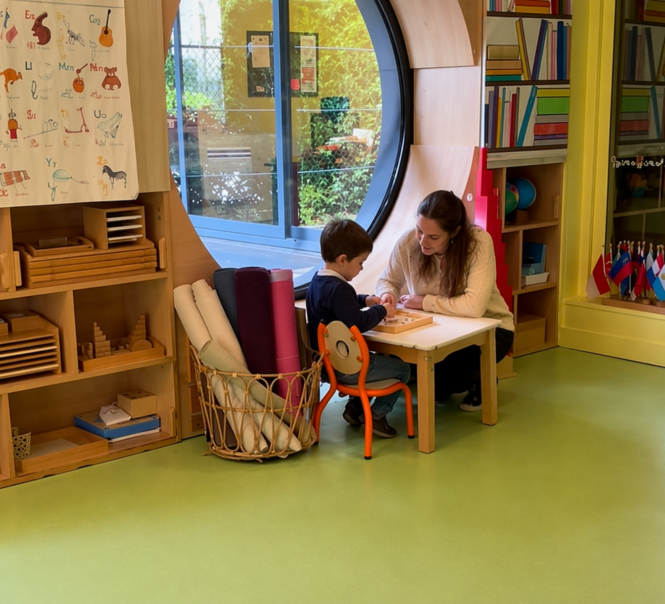 Enfant travaillant avec une éducatrice à une petite table dans une classe Montessori avec matériel en bois et grande fenêtre ronde